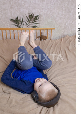 Boy in headphones listens to a meditation for children before going to bed. Concept of a healthy nervous system of children. Relaxation before sleep. Blond guy is relaxing at home. Hearing therapy Boy in headphones listens to a meditation for children before going to bed. Concept of a healthy nervous system of children. Relaxation before sleep. Blond guy is relaxing at home. Hearing therapy 86969380