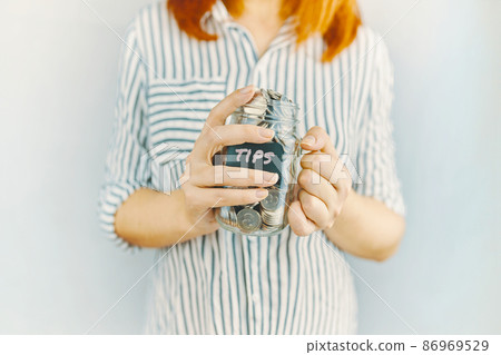 Glass bottle with silver coins in women's hands. Sticker with inscription on money jar. Redhead girl n white and black striped shirt holds her tips pot. 86969529