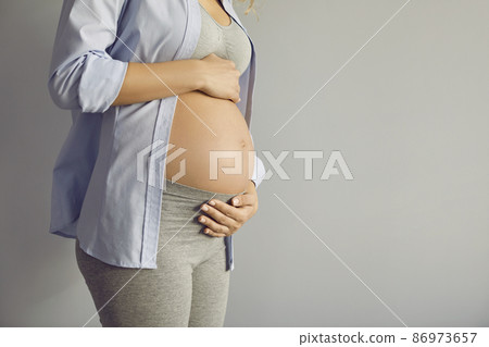 Close up of beautiful belly of pregnant woman on ninth month standing on gray background. Cropped image of mother-to-be in top , leggings and shirt holding both hands on her bare belly. Copy space. 86973657