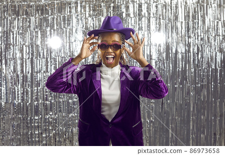 Happy dark-skinned young woman in velvet jacket suit arranges party on shiny disco background. Woman in purple clothes with funny facial expression wears party glasses standing on silver background. Happy dark-skinned young woman in velvet jacket suit arranges party on shiny disco background. Woman in purple clothes with funny facial expression wears party glasses standing on silver background. 86973658