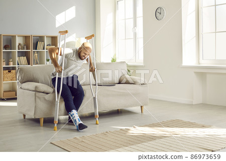 Home rehabilitation and recovery after physical injuries.Man with leg injury feels pain and difficulty trying to get up from sofa on crutches.Man wears bandage on his ankle and has difficulty walking Home rehabilitation and recovery after physical injuries.Man with leg injury feels pain and difficulty trying to get up from sofa on crutches.Man wears bandage on his ankle and has difficulty walking 86973659
