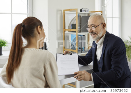 Friendly loan broker or estate agent meeting a client in the office. Smiling senior insurance agent or bank employee offers his customer a deal and shows documents with the contract agreement 86973662