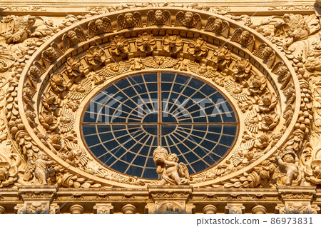 Lecce, Puglia, Italy. August 2021. The church of Santa Croce is the finest example of Lecce baroque. Detail of the facade illuminated by the warm light of the evening. The central rose window. 86973831