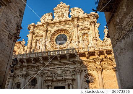 Lecce,Puglia,Italy.August 2021. The church of Santa Croce is the finest example of  baroque. The facade illuminated by the warm light of the evening peeps out between two houses in the historic center 86973836