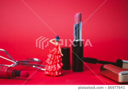 Decorative cosmetics products on red background - black mascara in gold tube, pink and red lipsticks, eyelash tongs. A toy woman in a red long dress with black hair. Female beauty and grooming concept Decorative cosmetics products on red background - black mascara in gold tube, pink and red lipsticks, eyelash tongs. A toy woman in a red long dress with black hair. Female beauty and grooming concept 86977641