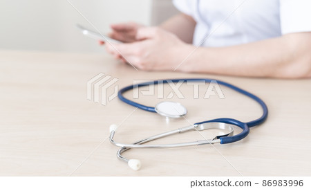 Unrecognizable female doctor sits at a desk and writes a message on a smartphone. Practitioner holds a phone in his hands and answers patients Unrecognizable female doctor sits at a desk and writes a message on a smartphone. Practitioner holds a phone in his hands and answers patients 86983996