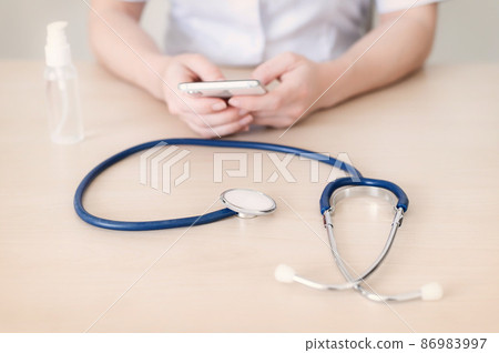 The doctor is writing on the smartphone during the break. Female cardiologist gives online consultation by cell phone. Stethoscope and hand antiseptic on the desktop. 86983997