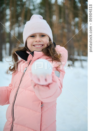Caucasian 4 years old cute girl in warm pink pastel down jacket showing a snowball, smiling looking at camera while enjoying active pastime outdoor on a snowy forest. Winter leisure activity concept 86984818