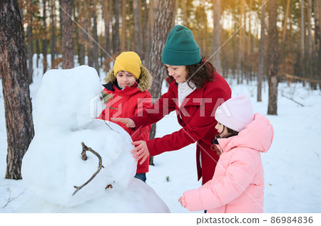 Young Caucasian happy family, mom, son and daughter are building a snowman in a snowy woodland, enjoying wonderful winter outdoor on a beautiful cool day with sunbeams falling through pine trees Young Caucasian happy family, mom, son and daughter are building a snowman in a snowy woodland, enjoying wonderful winter outdoor on a beautiful cool day with sunbeams falling through pine trees 86984836