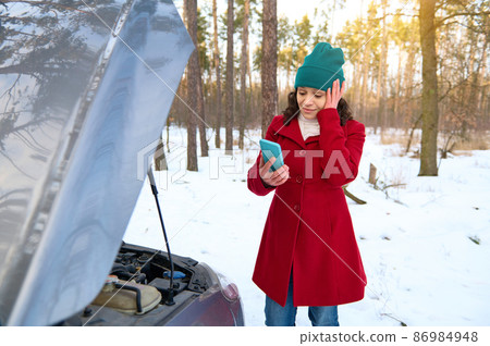 Stunned female driver with hand on temple calling tow truck or roadside assistance while standing in front of car with open hood on snow covered road. Car breakdown while traveling and car insurance Stunned female driver with hand on temple calling tow truck or roadside assistance while standing in front of car with open hood on snow covered road. Car breakdown while traveling and car insurance 86984948