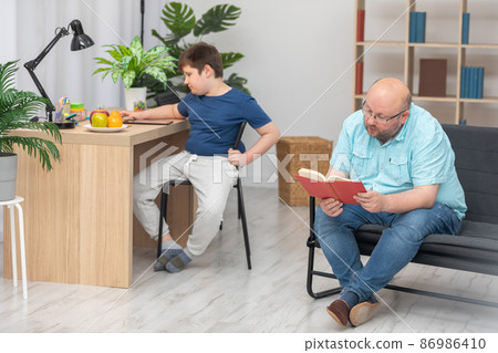 Dad is sitting on the sofa reading a book to his son and he is sitting at his desk. Dad is sitting on the sofa reading a book to his son and he is sitting at his desk. 86986410