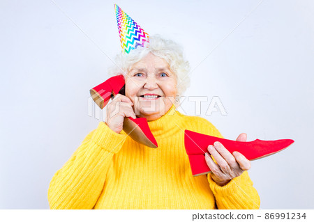 older woman with a gift wear yellow sweater and horn cap on a white background holding red high heels shoes 86991234