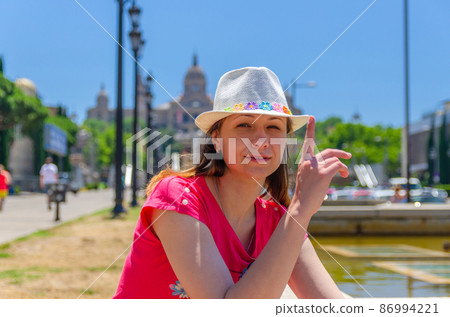Young woman traveler with red dress and hat is sitting near fountain in Barcelona in sunny summer day Young woman traveler with red dress and hat is sitting near fountain in Barcelona in sunny summer day 86994221