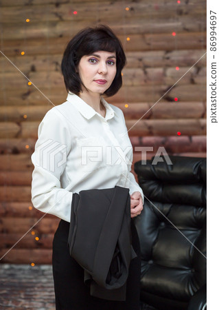 A brunette woman in a white blouse with a black jacket on her arm against a wooden wall with a garland A brunette woman in a white blouse with a black jacket on her arm against a wooden wall with a garland 86994697