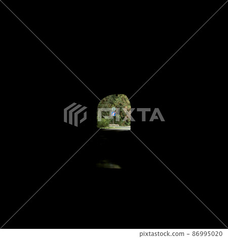 A road sign with a crossed-out arrow on the wooded roadside can be seen in the distance. View from dark long tunnel. low key 86995020