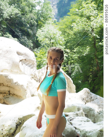 A girl in a blue swimsuit stands on a rocky cliff surrounded by a green grove 86995616