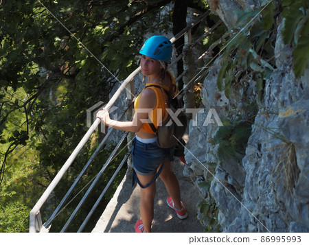 A girl in a helmet stands holding on to the railing on a narrow path of a rocky cliff in a green grove A girl in a helmet stands holding on to the railing on a narrow path of a rocky cliff in a green grove 86995993