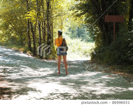 Girl tourist stands on a forked concrete road leading along a shady deciduous forest. Blurred image 86996031