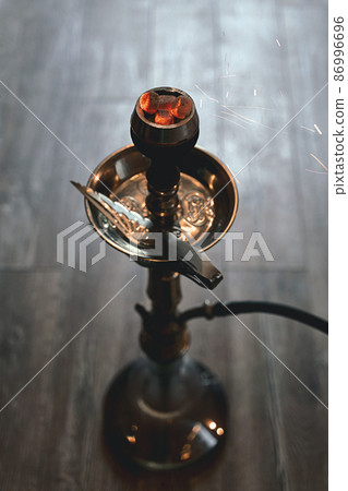Shisha hookah bowl with red hot coals and craft tobacco. Sparks from breathe. Modern hookah with coconut charcoal for relax and smoke. Beautiful sparks in long exposure on lounge floor background. Shisha hookah bowl with red hot coals and craft tobacco. Sparks from breathe. Modern hookah with coconut charcoal for relax and smoke. Beautiful sparks in long exposure on lounge floor background. 86996696