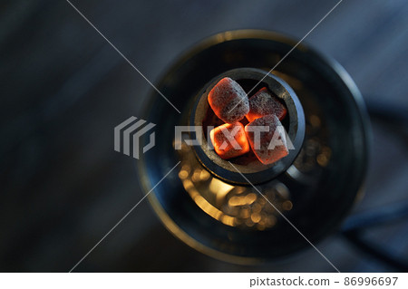 Shisha hookah bowl with red hot coals and craft tobacco. Modern hookah with coconut charcoal for relax and smoke. Beautiful bokeh and cozy dark lounge background. Top view. 86996697