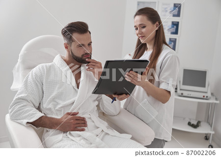 Administrator shows clipboard with form to client sitting in armchair in cosmetological clinic Administrator shows clipboard with form to client sitting in armchair in cosmetological clinic 87002206