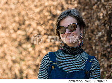 Portrait of a thirty year old white smiling lady with sunglasses with a background of a brown buxus in a Brussels city park. 87002548