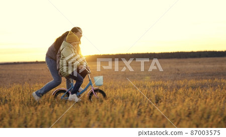 childhood dream. woman mother teaches child to ride bike in the park. Driving lesson for kid from mom at sunset. parent helps child to keep balance and pedal. Kid learns steer bike sun. happy family 87003575