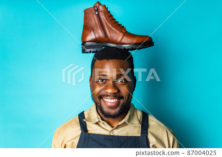 latin hispanic man in black apron showing brown leather shoes in blue studio background 87004025