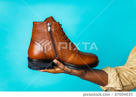 latin hispanic man in black apron showing brown leather shoes in blue studio background 87004035