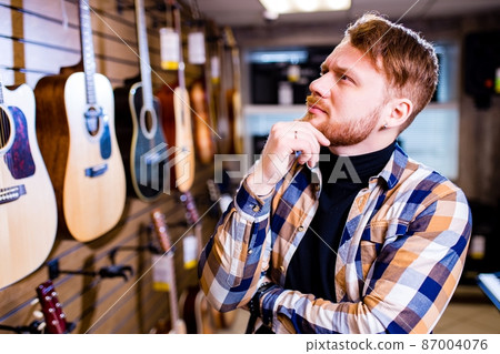 redhaired bearded handsome man in brown plaid casual shirt choosing a guitar in a music store 87004076
