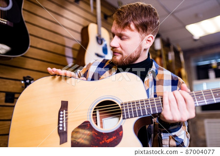 redhair ginger man is choosing quality acoustic guitar in guitar shop 87004077