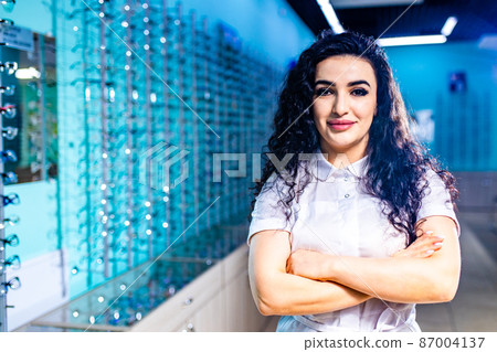 eastern ethnic woman in white medical coat robe selling glasses in optical store 87004137