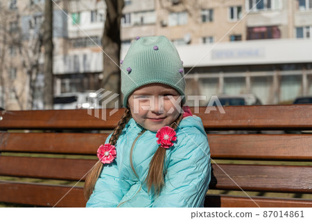 Portrait of a 5-year-old girl sitting on a park bench. Shy girl with long hair braided into pigtails. A child in a blue jacket and hat. Springtime. Portrait of a 5-year-old girl sitting on a park bench. Shy girl with long hair braided into pigtails. A child in a blue jacket and hat. Springtime. 87014861