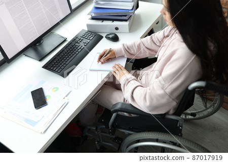 Disabled woman in wheelchair with documents uses computer Disabled woman in wheelchair with documents uses computer 87017919
