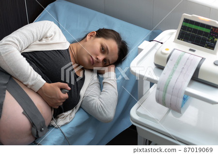 Pregnant Woman On Hospital Couch During Medical Control Cardiotocography. Gynecologist Doctor Hands Turn On Adjust CTG Device Clinic Consultant. Examine Expectant Belly Baby Mother Healthcare Check Up Pregnant Woman On Hospital Couch During Medical Control Cardiotocography. Gynecologist Doctor Hands Turn On Adjust CTG Device Clinic Consultant. Examine Expectant Belly Baby Mother Healthcare Check Up 87019069