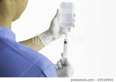 Hands of a doctor or nurse wearing rubber gloves and using a syringe to suck Sodium Chloride in a clinic or hospital alone with a white background. Hands of a doctor or nurse wearing rubber gloves and using a syringe to suck Sodium Chloride in a clinic or hospital alone with a white background. 87019930
