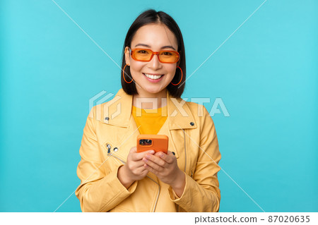 Smiling asian girl in sunglasses, using smartphone app, holding mobile phone, standing over blue background Smiling asian girl in sunglasses, using smartphone app, holding mobile phone, standing over blue background 87020635