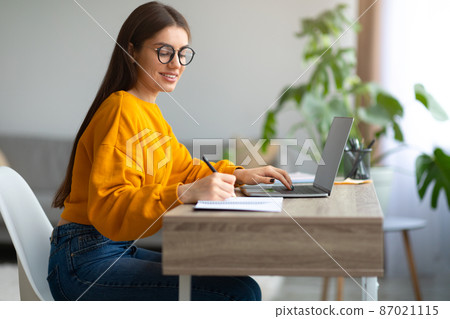 Smiling young woman in glasses sitting at desk, working on laptop, writing in notebook at home, free space Smiling young woman in glasses sitting at desk, working on laptop, writing in notebook at home, free space 87021115