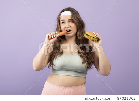 Depressed plus size european millennial female in sportswear eats carrot and holds burger Depressed plus size european millennial female in sportswear eats carrot and holds burger 87021302