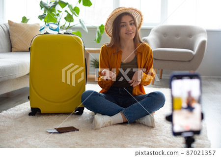 Young travel vlogger recording video content on smartphone, sitting on floor near suitcase, speaking to blog followers 87021357