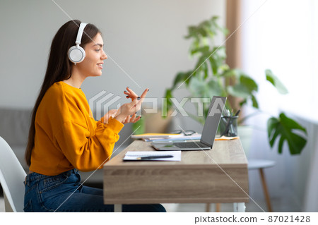 Happy young Caucasian female wearing headphones, using laptop, working or studying online from home, empty space 87021428