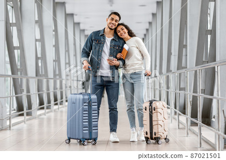 Cheerful Young Middle Eastern Couple Standing At Airport With Luggage And Smartphone Cheerful Young Middle Eastern Couple Standing At Airport With Luggage And Smartphone 87021501