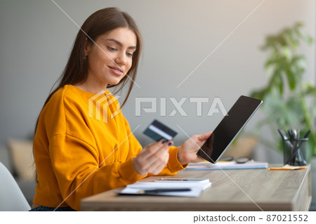 Cheerful young lady with tablet pc and credit card making online order, using remote banking service at home Cheerful young lady with tablet pc and credit card making online order, using remote banking service at home 87021552