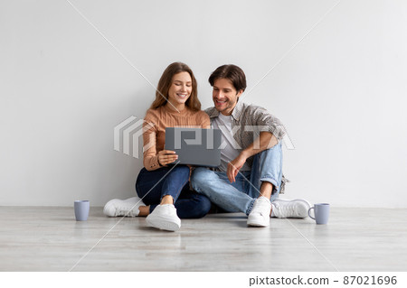 Glad smiling young european man and lady look at laptop planning new interior, sitting on floor in empty room 87021696