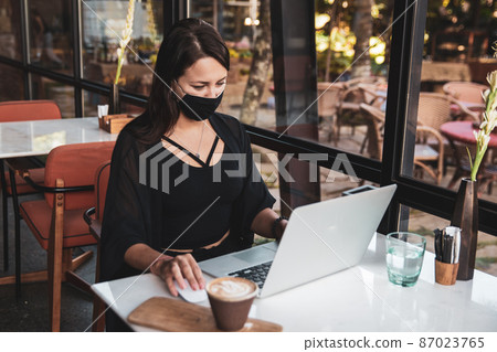 Young woman freelancer with protective medical black mask on face working with laptop in cafe Young woman freelancer with protective medical black mask on face working with laptop in cafe 87023765
