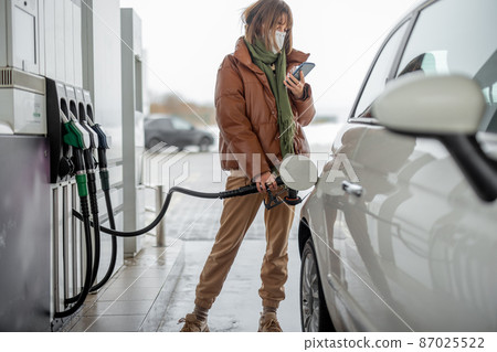 Woman in face mask refueling car with a gasoline using smart phone Woman in face mask refueling car with a gasoline using smart phone 87025522