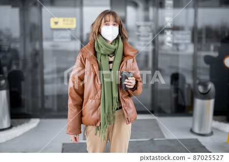 Woman in mask going out from shop with a coffee cup Woman in mask going out from shop with a coffee cup 87025527