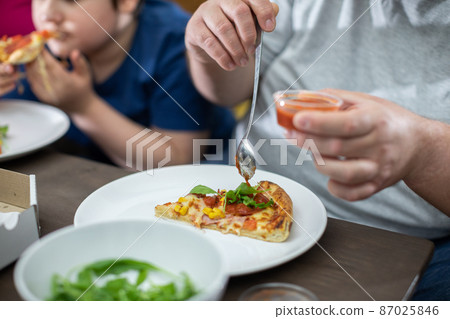 Father is putting tomato sauce on a slice of pizza. Mother waits for her turn. Father is putting tomato sauce on a slice of pizza. Mother waits for her turn. 87025846