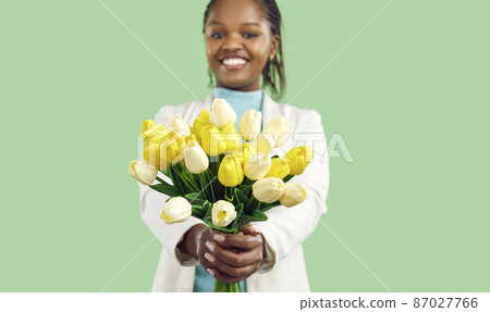Happy Afro American girl holding a bunch of spring flowers and smiling. Closeup studio shot of a beautiful bouquet of fresh yellow and white tulips in the hands of a pretty young black woman 87027766