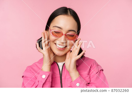 Close up of stylish korean girl in sunglasses, smiling happy, posing against pink background. People face concept Close up of stylish korean girl in sunglasses, smiling happy, posing against pink background. People face concept 87032260
